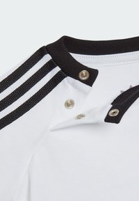 Camicia bianca con colletto e rifiniture nere, caratterizzata da strisce bianche e nere sulle maniche. Chiusure a scatto sul colletto aggiungono un tocco di dettaglio.