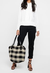 Frau trägt eine weiße Bluse, schwarze Hose und schwarze Riemchensandalen und hält eine große schwarz-beige karierte Tragetasche.