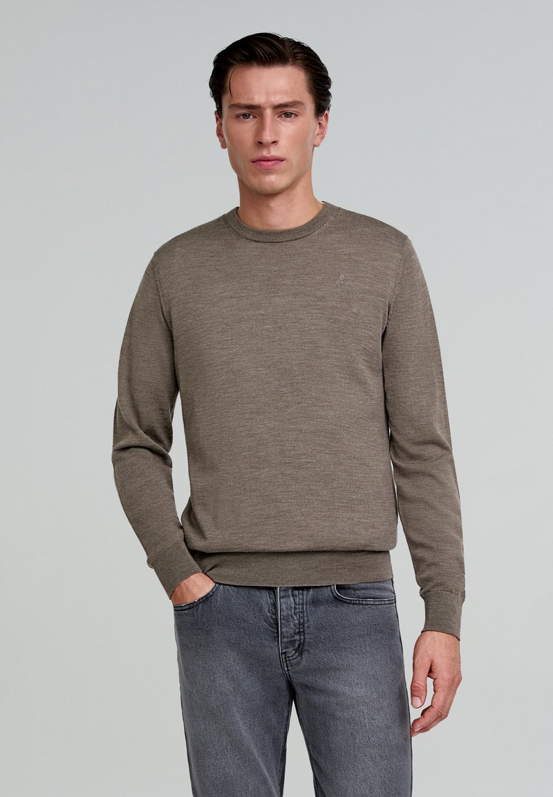 Brauner Strickpullover mit rundem Halsausschnitt und gerippten Bündchen, kombiniert mit grauen Jeans. Dezentes Logo-Detail auf der Brust. Glatte Textur.