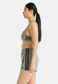 Žena v bočním profilu, oblečená v olivově zeleném sportovním brá a šortkách s černými pruhy, stojící před jednoduchým bílým pozadím.