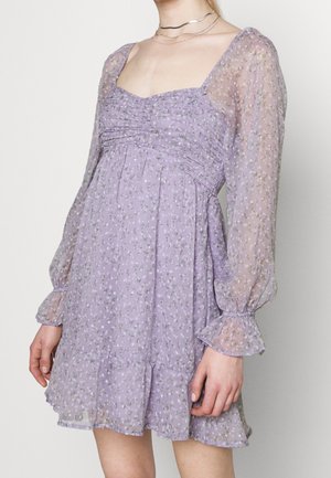 Femme portant une mini-robe lilas avec des manches longues bouffantes transparentes et un petit imprimé floral, associée à des colliers argentés superposés.