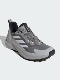 Zapatilla de trail running ligera en gris, con parte superior de malla texturada, franjas de acento blancas, puntera reforzada y suela negra rugosa.