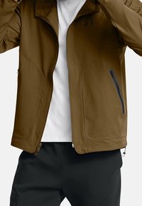 Brun jacka med en mjuk yta, dragkedjeförsedda fickor och ståkrage. Har kontrasterande svarta detaljer och två sidodragkedjor.