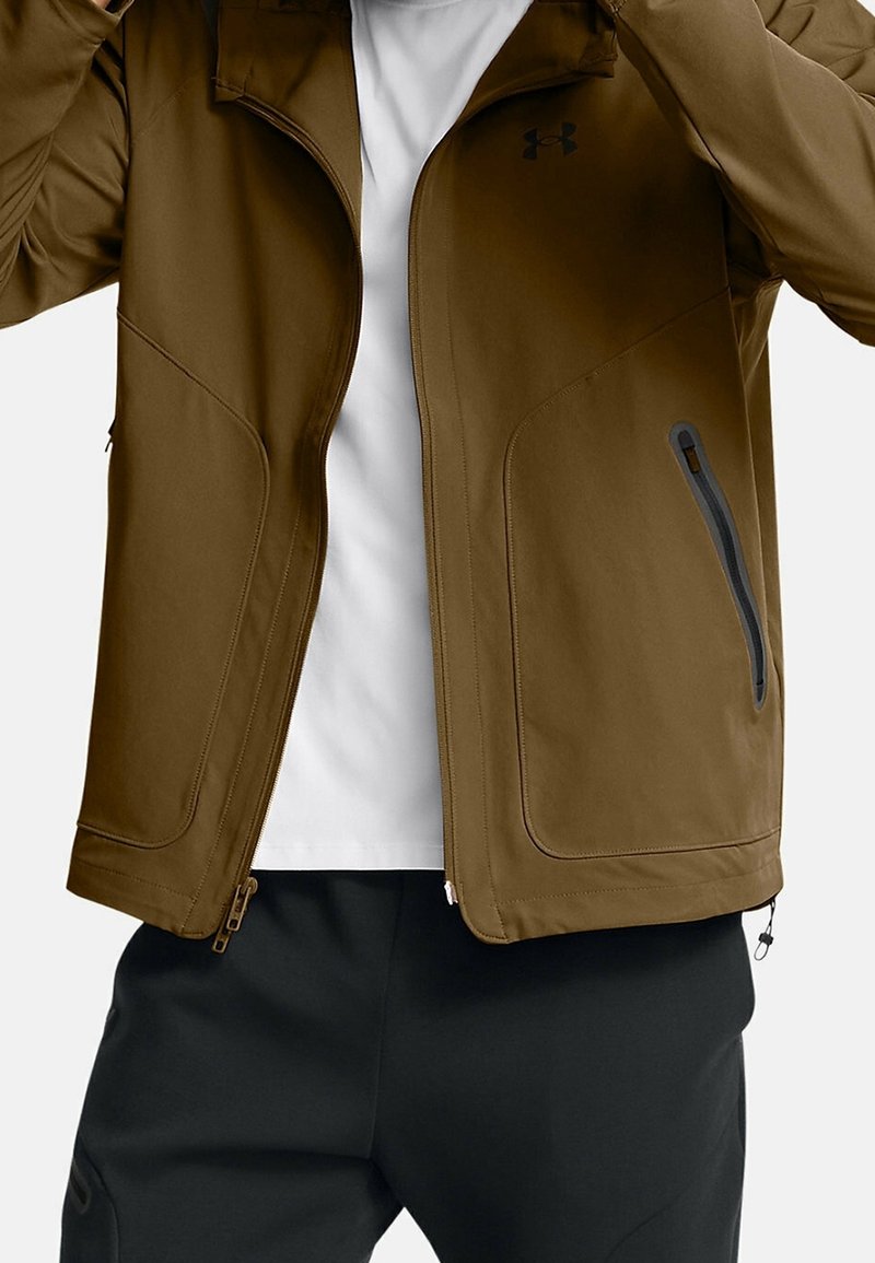 Brun jacka med en mjuk yta, dragkedjeförsedda fickor och ståkrage. Har kontrasterande svarta detaljer och två sidodragkedjor.