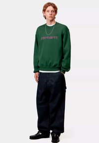 Sudadera verde con el logo "carhartt." en púrpura, combinada con jeans oscuros de corte holgado y zapatos negros. Diseño sencillo, ajuste casual.