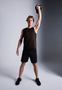 Kettlebell preta segurada acima da cabeça por uma pessoa com uma blusa sem mangas castanha e calções pretos, com cabelo curto e corpo atlético, contra um fundo degradê.