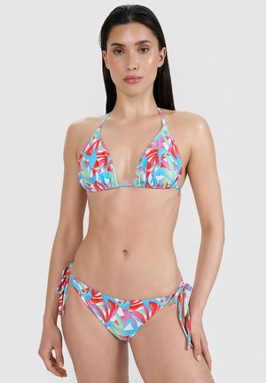 Kobieta ubrana w kolorowe stringi bikini w tropikalny wzór z wiązaniami po bokach i wiązaniem na szyi, na jednolitym jasnym tle.
