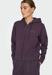 Sieviete, kurai mugurā ir tumši purpura krāsu zip-hoodie un atbilstošas bikses, ar rokām kabatās, skatās pa kreisi uz viendabīgas krāsas fona.