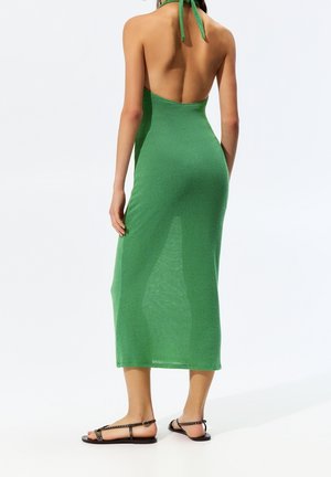 Vihreä halterikaulainen mekko, jossa istuva yläosa ja avoin selkä, polven pituinen, valmistettu kevyestä, teksturoidusta kankaasta. Minimalistiset mustat sandaalit.