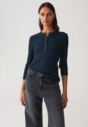 Vrouw in een marineblauw geribbeld langemouwtop met knoopsluiting en vervaagde zwarte hoge taille wijdpijpen jeans tegen een effen achtergrond.