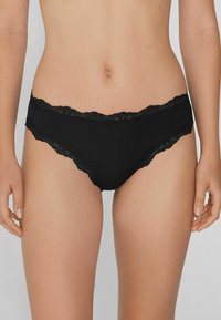 Mutande da donna a vita bassa con pizzo nero, indossate da una persona in piedi con le braccia rilassate ai lati.