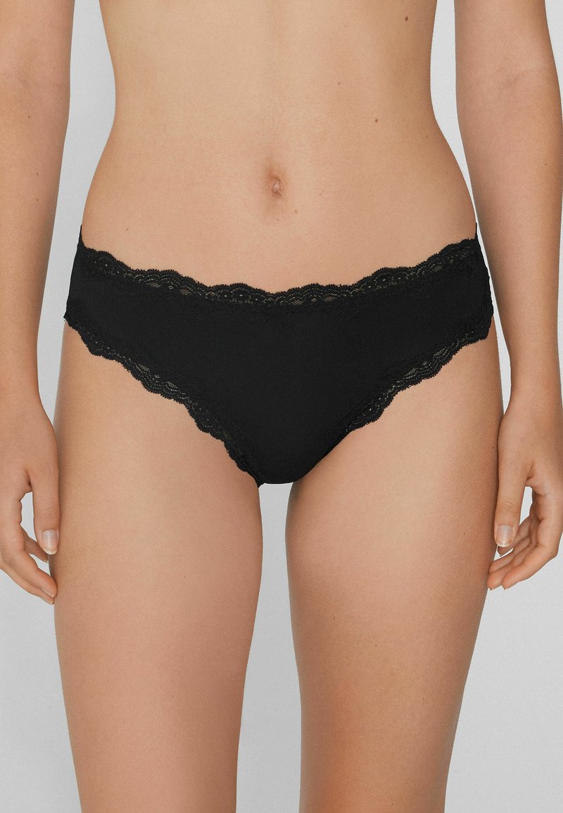 Mutande da donna a vita bassa con pizzo nero, indossate da una persona in piedi con le braccia rilassate ai lati.