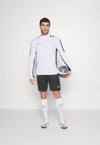 Cămașă sport albă cu mâneci lungi și accente negre, asortată cu pantaloni scurți negri, șosete albe și ghete de fotbal colorate. Minge de fotbal ținută în mână.