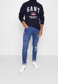 Marineblauer Pullover mit fettgedrucktem weißen Text und Sternen, kombiniert mit ausgebleichten blauen Jeans mit einer geflickten Tasche und weißen Sneakers mit roten Akzenten.
