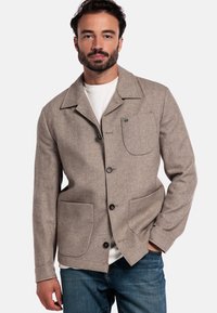 Bruine wollen blend jas met een kraag, knopen aan de voorkant, twee grote zakken en subtiele textuurdetails, gedragen over een wit shirt.