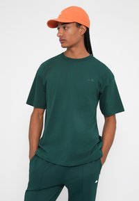 Mørkegrønn bomull t-skjorte med avslappet passform, rund hals og korte ermer; kombinert med teal bukser. Oransje baseballcaps brukt på hodet.