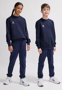 Completo sportivo di felpa e pantaloni in blu navy, con un logo sul petto, polsini elastici e sneaker di colore neutro.