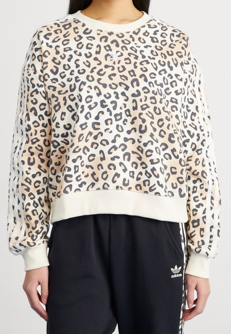 Frau trägt einen beigefarbenen und schwarzen Leopardenmuster-Adidas-Pullover mit weißen Streifen an den Ärmeln sowie schwarze Adidas-Hose mit Logo auf dem linken Oberschenkel.
