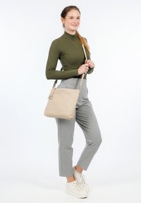 Borsa a tracolla in pelle beige con finitura texturizzata, tracolla regolabile e un accento in metallo circolare. Indossata con pantaloni grigi a vita alta e sneakers bianche.