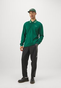 Polo verde de manga larga con cuello a rayas, logo bordado, combinado con pantalones negros con rayas blancas y zapatos negros de cordones.