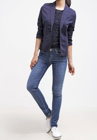 Femme en blazer bleu marine foncé, top en dentelle noire, jean slim bleu et baskets blanches, se tenant avec une jambe croisée devant l'autre.