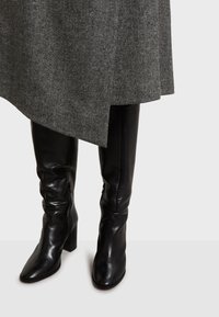 Crne kožne čizme do koljena s debelim blok-petom, s sjajnim finišem i bočnim zatvaračem za lako oblačenje. Visible su suknja s uzorkom riblje kosti.