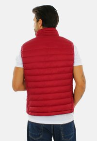 Gilet imbottito rosso con colletto alto, caratterizzato da sezioni a righe orizzontali e una texture liscia, indossato sopra una camicia bianca.