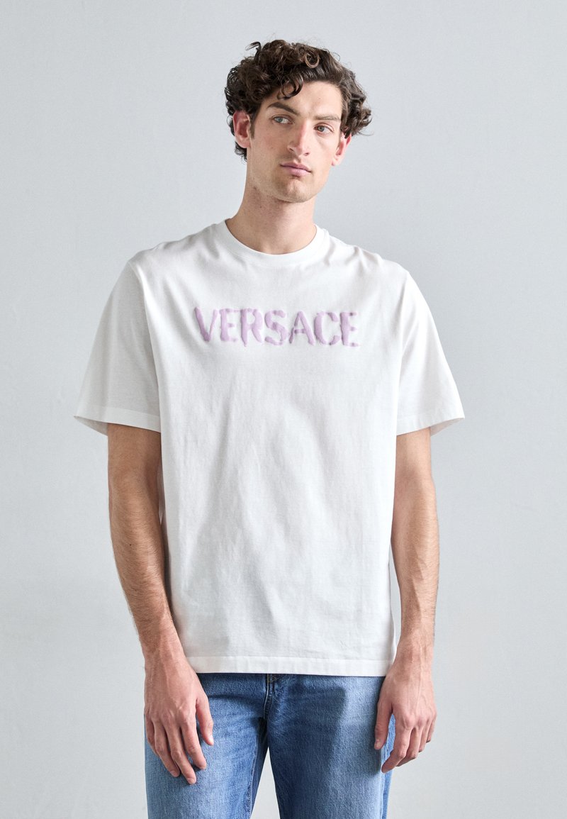 Balta medvilnė marškinėliai su tekstūruotu rožiniu siuvinėjimu "VERSACE" logotipu krūtinėje; trumpomis rankovėmis ir laisvu siluetu.