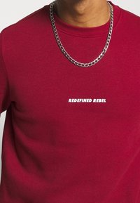 Sweat-shirt rouge avec encolure côtelée portant le texte blanc "REDEFINED REBEL". Un collier chaîne argenté sert d'accessoire sur le tissu.