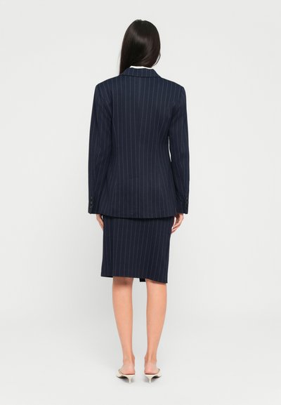 Kvinne med langt mørkt hår som har på seg en marineblå pinstripe-blazer og matchende skjørt til kneet, står med ryggen mot kameraet på en hvit bakgrunn.