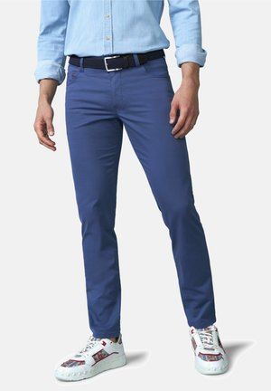 Mann trägt blaue, schmal geschnittene Hose, hellblaues Hemd mit aufgekrempelten Ärmeln, schwarzen Gürtel und weiße Sneakers mit roten und gemusterten Details.