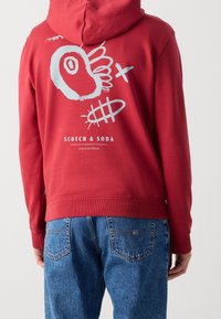 Felpa rossa con cappuccio, caratterizzata da un design grafico che presenta un occhio stilizzato e dettagli in azzurro chiaro. Include il testo del logo al centro. Tessuto misto cotone.