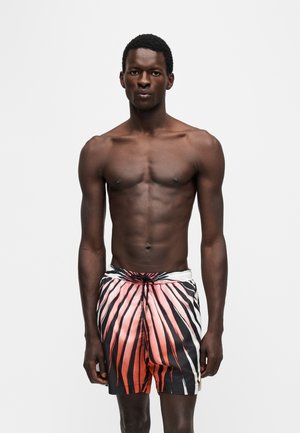Mannelijk model met een donkere huid, dragend oranje en zwarte gestreepte zwembroek, staand tegen een effen witte achtergrond.