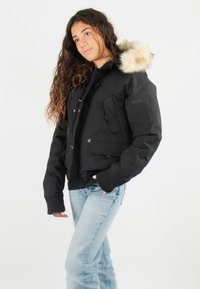 Veste noire isolée avec une capuche bordée de fourrure, dotée de poches multiples et de poignets côtelés, associée à un jean bleu clair.