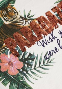 Vit t-shirt med en färgglad tiger grafik, gröna blad, rosa blommor och glänsande rosaguld paljetttext som säger "PARADISE."