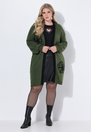 Cardigan en tricot surdimensionné de couleur vert foncé avec des broderies florales noires, à devant ouvert et aux poignets côtelés, porté sur un haut noir et une jupe en cuir.