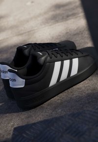 Baskets Adidas noires avec des bandes blanches et une languette au talon, placées côte à côte sur une surface en béton texturée avec un éclairage ombragé.