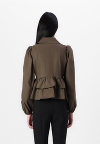Veste peplum marron avec des manches bouffantes, un col saillant et un ourlet en volants superposés. Présente une texture de tissu lisse et une coupe ajustée.