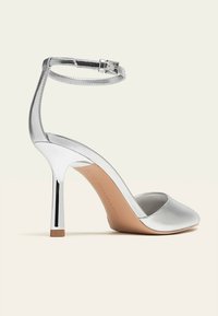 Stříbrná vysoce podpatková bota s špičatou špičkou, tenkým řemínkem kolem kotníku s přezkou a štíhlým jehlovým podpatkem na neutrálním pozadí.