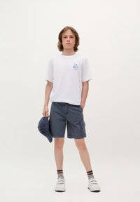 T-shirt bianca in cotone con grafica blu, pantaloni cargo scuri, cappellino blu e sneakers bianche con accenti neri. Calze a righe visibili.