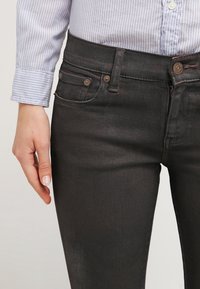 Jeans in denim nero con un design fitted, tasche anteriori con dettagli metallici e una texture liscia. Indossati con una camicia a righe blu.