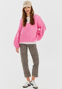 Różowy, dziany sweter, biały krótki top, spodnie typu cropped w leopardzi wzór oraz białe sneakersy z czarnymi akcentami. Czapka z logo dopełnia całość.