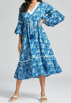 Kvinde iført en blå midi-kjole med blomsterprint og klokkeærmer, med hvid kvastbånd i taljen og cremefarvede sandaler, stående med den ene hånd på hoften.