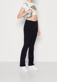 Uppflutna corduroy-jeans i mörk marinblå, kombinerat med en vit kortärmad topp med vågig nederkant som har en katt- och fjärilsdesign. Tillbehör inkluderar en metallisk handväska.