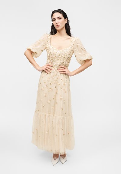 Femme portant une robe midi beige, transparente, brodée de fleurs, avec des manches bouffantes, debout les mains sur les hanches et des chaussures blanches pointues.