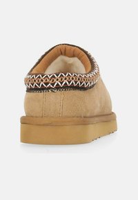 Tan ruskindslipper med rund tå, tekstureret brun og hvid vævet trim rundt om kraven, og en lys brun gummisål.