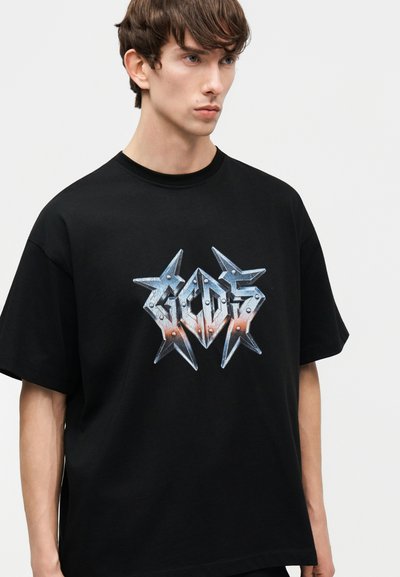 Νεαρός άνδρας με κοντά καστανά μαλλιά που φοράει μια μαύρη φαρδιά μπλούζα με μεταλλικό λογότυπο με αγκάθια σε αποχρώσεις μπλε και πορτοκαλί.