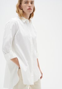 Balta kurpju jebša ar apkakli un sarullētām piedurknēm, ar brīvu piegriezumu un garāku aizmuguri, izgatavota no gludas, vieglas auduma.