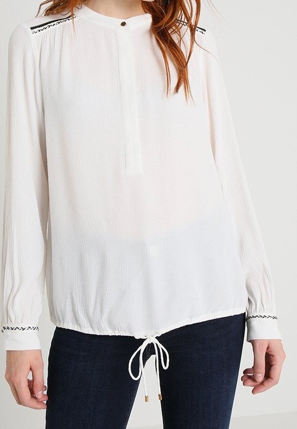 Blouse blanche à manches longues avec un tissu texturé, un col rond et un cordon à l'ourlet. Présente un accent noir sur les coutures des épaules.