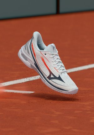 Chaussure de tennis blanche et rouge en l'air au-dessus d'un court en terre battue, avec de la poussière d'argile visible autour de la semelle et une ligne de terrain de tennis en arrière-plan.